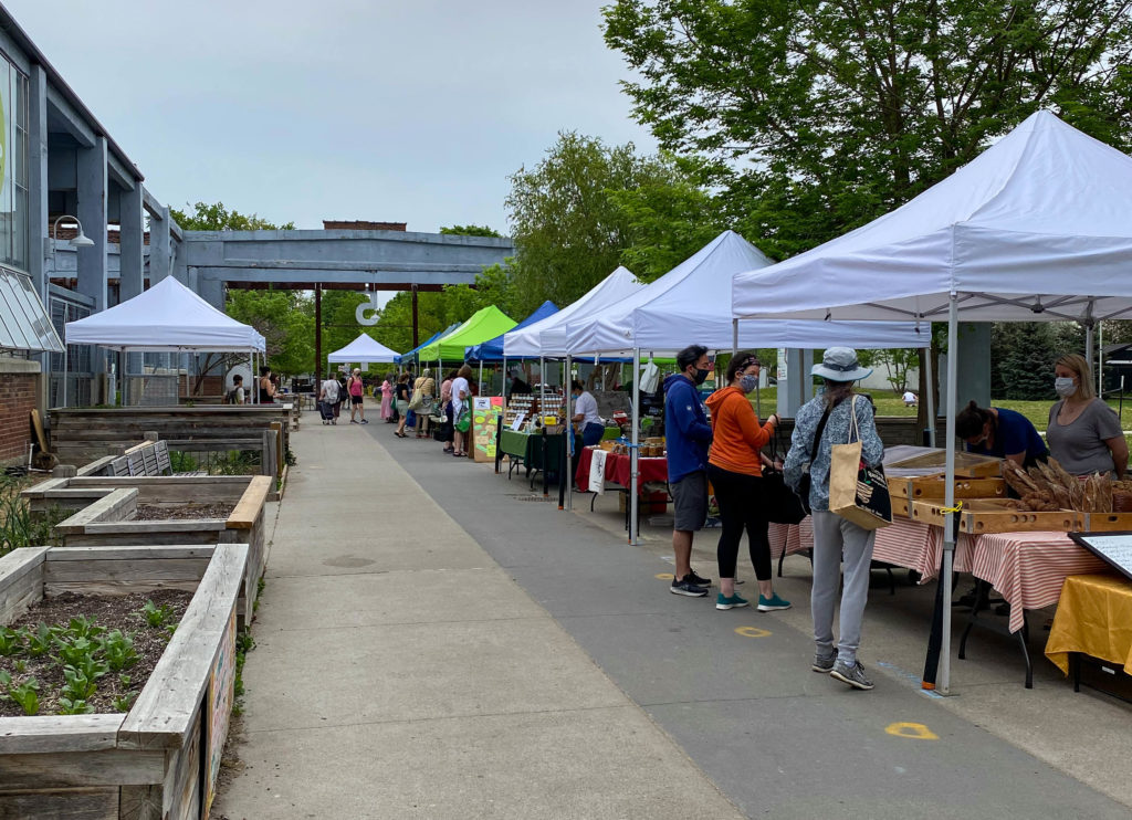 The sun is shining but it is a heavily clouded sky. Both images depict rows of outdoor event tents where vendors are situated, all lined up next to each other. Market goers can be seen at stalls or on the main walkway, but it is relatively sparse.