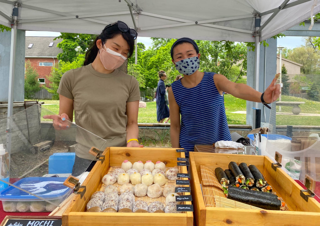 At a vendor stand, two Asian women stand presenting their food for sale in large wooden display cases. The left container is full of rows of mochi, and the right has stacks of long sushi rolls.