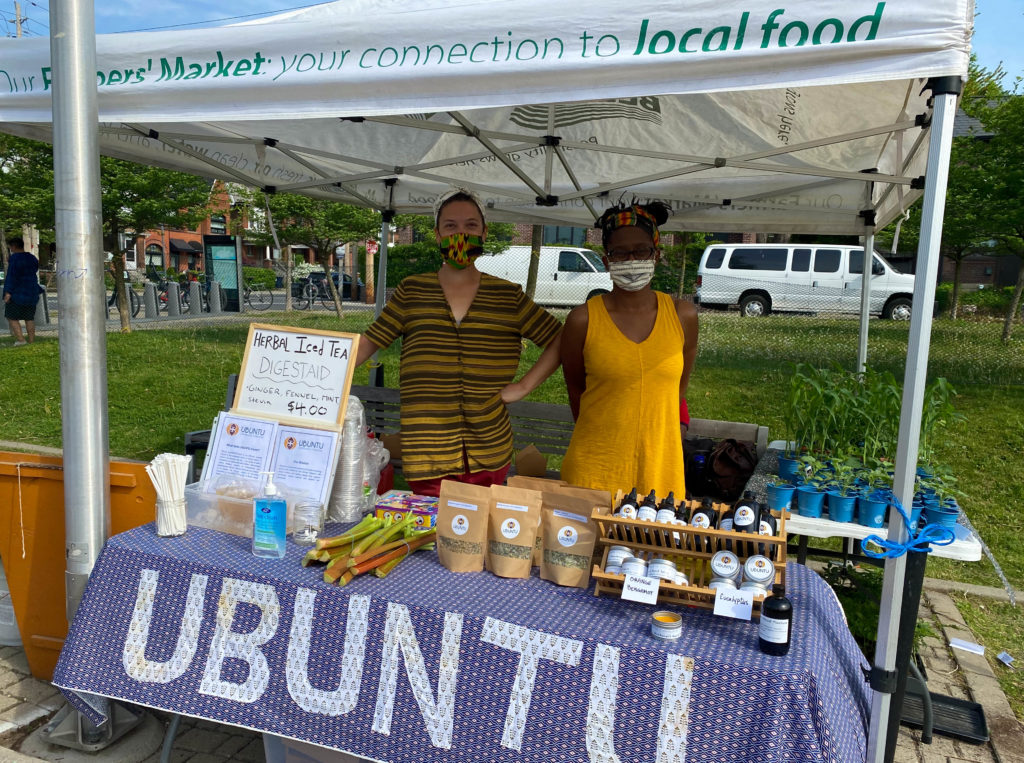 At a vendor stand, two darker-skinned women stand selling teas, plant seedlings, some vegetables, and some bottled products. The tablecloth has "UBUNTU" on the front in large capitalized letters.