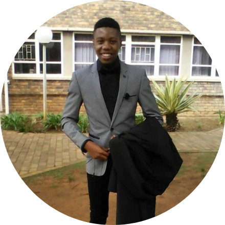 Sethu's photo: Picture of South African person wearing a grey blazer in front of a sidewalk.